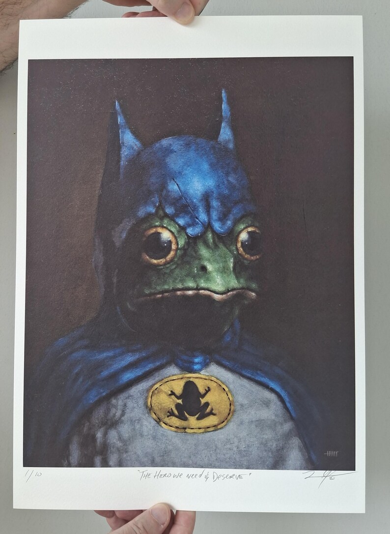 May include: A print featuring a frog dressed as Batman, with a blue cowl and cape. The frog has large, expressive eyes and a green face. A yellow oval emblem with a frog silhouette is on the chest. The text "The Hero we need & Deserve" is at the bottom.