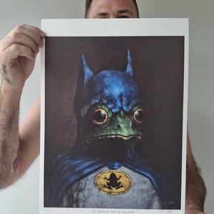 May include: A print featuring a frog dressed as Batman, with a blue cowl and cape, and a yellow emblem. The frog has large, expressive eyes and a green face. The print includes the text "The Heroes we need & Deserve."