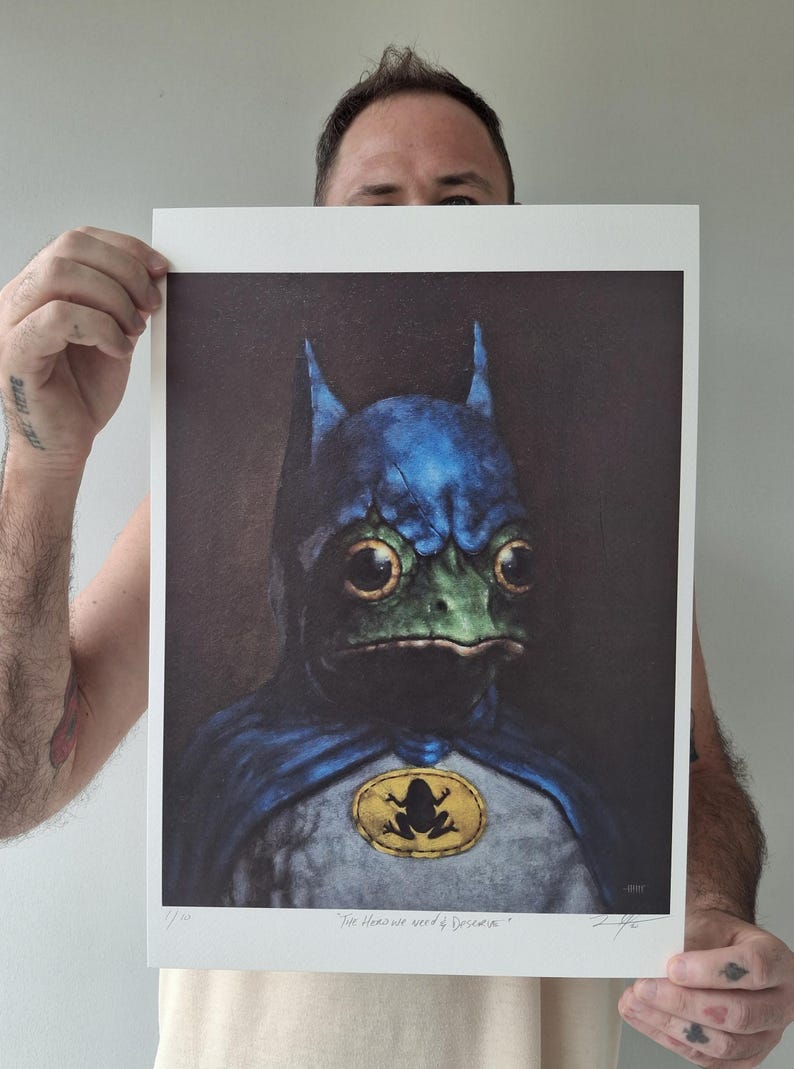 May include: A print featuring a frog dressed as Batman, with a blue cowl and cape, and a yellow emblem. The frog has large, expressive eyes and a green face. The print includes the text "The Heroes we need & Deserve."