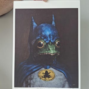 May include: A print featuring a frog dressed as Batman, with a blue cowl and cape. The frog's face is green, and it has large, expressive eyes. A yellow emblem with a frog silhouette is on the chest. The text "The Hero We Need & Deserve" is at the bottom.