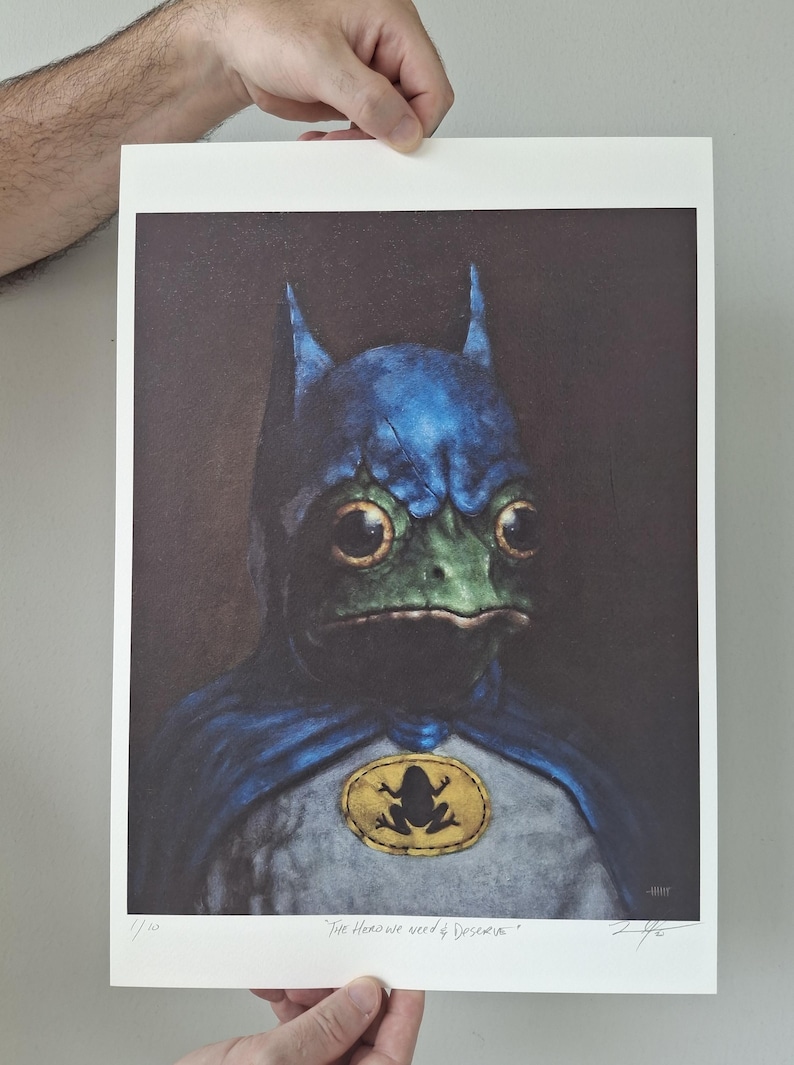 May include: A print featuring a frog dressed as Batman, with a blue cowl and cape. The frog's face is green, and it has large, expressive eyes. A yellow emblem with a frog silhouette is on the chest. The text "The Hero We Need & Deserve" is at the bottom.