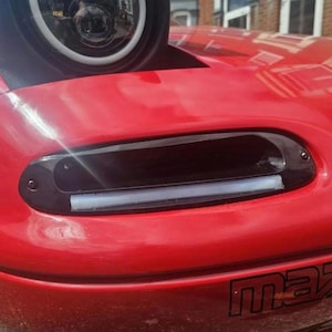 May include: A bright red car front end featuring a pop-up headlight with a circular light assembly and a black air intake vent integrated into the bonnet. The vent contains a horizontal white LED light strip and is secured with visible black screws.
