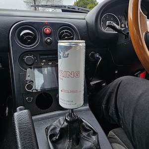 May include: A white ceramic mug with a black handle and the text "But First, Coffee" in a bold, black font. The mug is set against a blurred background. The mug appears to be approximately 10 cm tall.