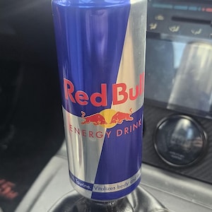 May include: A Red Bull energy drink can, predominantly blue and silver, is positioned on a black gear stick inside a car. The can displays the Red Bull logo and the words "ENERGY DRINK".