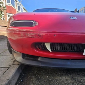 May include: A red Mazda Miata sports car with a black front bumper and a black grill. The car has a Mazda logo on the side and white fangs attached to the front. The car is parked on a street.