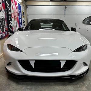 May include: A white sports car with a black front bumper and a black grille shaped like vampire fangs. The car is parked in a garage with a white door and a banner with Japanese text.