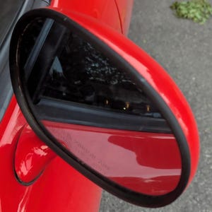 May include: A close-up shot of a red car's side mirror, outlined with a black trim. The mirror reflects the surroundings, showing a hint of the car's interior and exterior. The car's bodywork is a bright red.