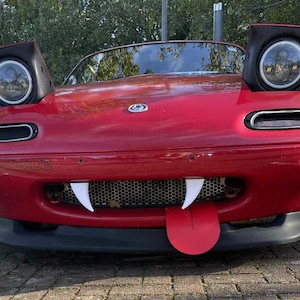 May include: A red Mazda Miata convertible with custom modifications. The car has pop-up headlights, a black front bumper, and a grille with white fangs and a red tongue. The car is parked on a brick surface.