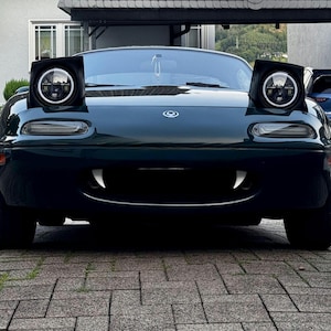 May include: Dark green Mazda Miata sports car with custom LED headlights. The car has a unique design with white fangs on the front bumper. The car is parked on a brick driveway.