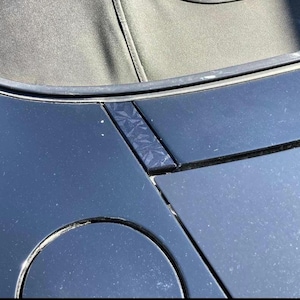 May include: A close-up view of a black car's exterior, focusing on the convertible top's edge and the bodywork. The paintwork has a glossy sheen, with a textured detail on a small section. The top is a dark grey fabric.