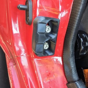 May include: A close-up of a car door interior. The door is red, with a black plastic component secured by screws. A black door handle and a black door latch are also visible. The interior trim is black.
