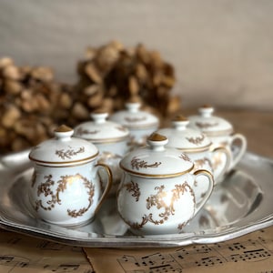 May include: A collection of six white porcelain sugar bowls with lids, each adorned with gold floral patterns and trim. The bowls are arranged on a silver tray, with musical notes in the foreground.