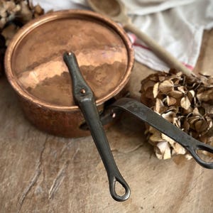 May include: A round copper pot with a lid and a long, dark metal handle. The lid features a matching dark metal crossbar handle secured with copper rivets. The exterior surface of the pot shows a hammered texture and a warm, metallic finish.