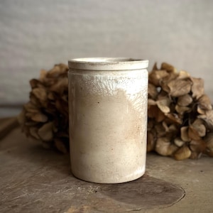 May include: A cylindrical, off-white ceramic container with a slightly textured surface. The container has a rounded top and a base. The background features dried, brown foliage. The container is likely a vintage or antique piece.