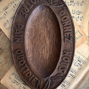 May include: An oval dark brown wooden bread bowl with a carved rim featuring wheat stalks and the French text NOTRE PAIN QUOTIDIEN DONNEZ NOUS. The bowl has a deep, smooth center and a visible wood grain texture throughout the entire carved surface.