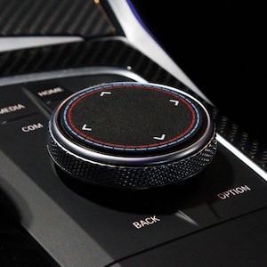 May include: A close-up of a black control knob with a silver ring and red and blue accents. The knob has four directional arrows and is labeled "COM", "MEDIA", "HEAR", "BACK", and "OPTION".