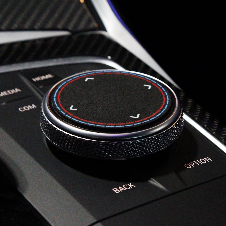 May include: A close-up of a black control knob with a silver ring and red and blue accents. The knob has four directional arrows and is labeled "COM", "MEDIA", "HEAR", "BACK", and "OPTION".