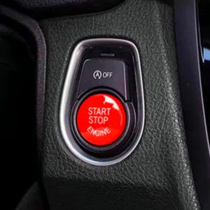May include: A close-up of a car's start-stop engine button. The button is red with white text that reads "Start Stop Engine". The button is surrounded by a silver ring and is located on a black dashboard.