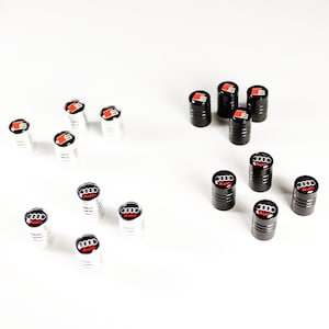May include: A set of 12 black and silver tire valve stem caps with the Audi logo. The caps are arranged in two rows, with the black caps on the top row and the silver caps on the bottom row.