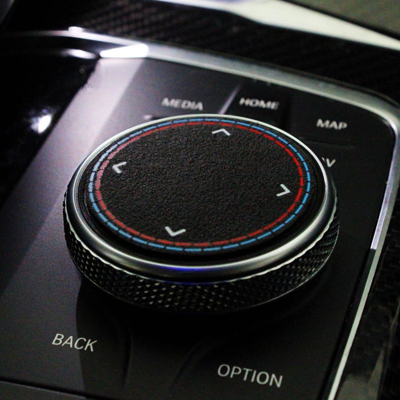 May include: A close-up of a black control knob with a silver ring and red and blue markings. The knob has four directional arrows, indicating up, down, left, and right. The knob is labeled "MEDIA", "HOME", "MAP", "BACK", and "OPTION".