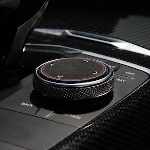 May include: A black knob with a silver ring and a red and blue ring around a black center. The knob has a textured surface and is labeled "NAV" and "MAP".