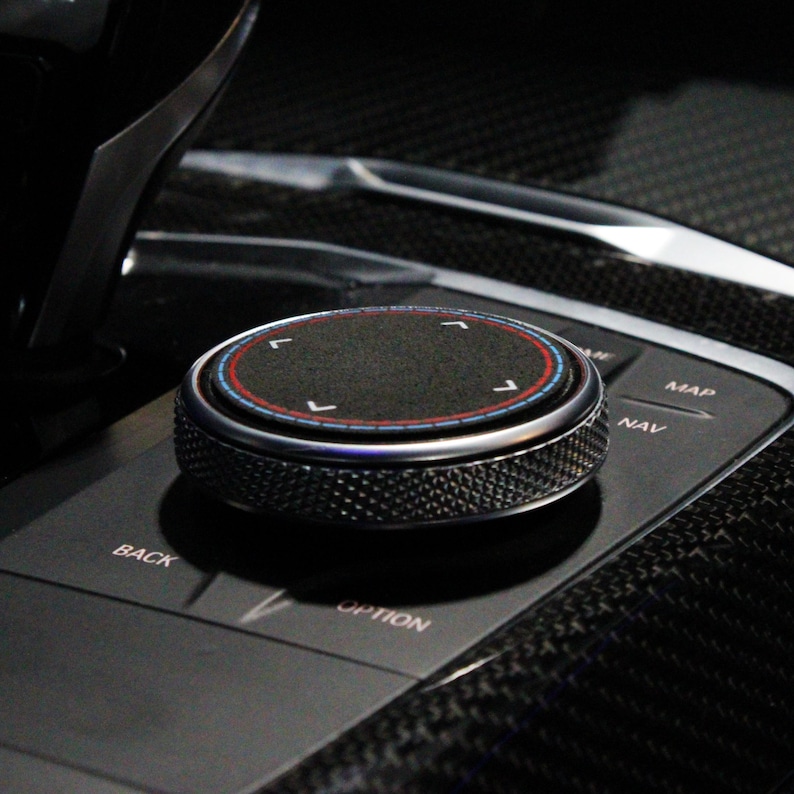 May include: A black knob with a silver ring and a red and blue ring around a black center. The knob has a textured surface and is labeled "NAV" and "MAP".
