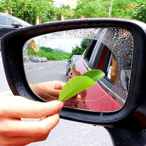 May include: A black car side mirror with a clear, water-repellent film applied. The mirror reflects a road scene with other vehicles and greenery. Two green leaves are held near the mirror's surface.