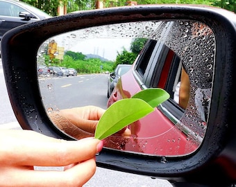 May include: A black car side mirror with a clear, water-repellent film applied. The mirror reflects a road scene with other vehicles and greenery. Two green leaves are held near the mirror's surface.