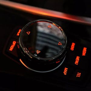 May include: A close-up of a black car's control panel with a round, silver knob and red illuminated buttons labeled "Option", "Radio", "Media", "Menu", "Tel", and "Map".