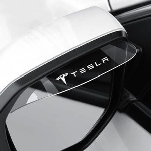 May include: A black side view mirror with a clear plastic rain guard. The rain guard has the word "TESLA" printed in white.