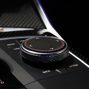 May include: A close-up of a black control knob with a silver ring and a textured surface. The knob has four directional arrows in a circle, with red and blue accents. The knob is labeled "COM", "MEDIA", "BACK", and "OPTION".