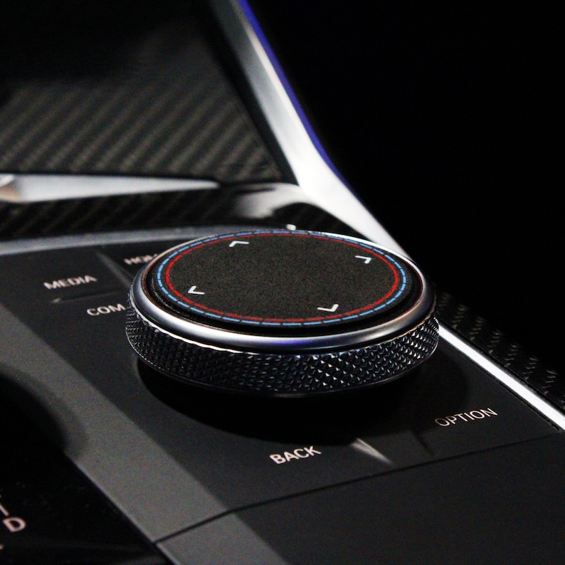 May include: A close-up of a black control knob with a silver ring and a textured surface. The knob has four directional arrows in a circle, with red and blue accents. The knob is labeled "COM", "MEDIA", "BACK", and "OPTION".