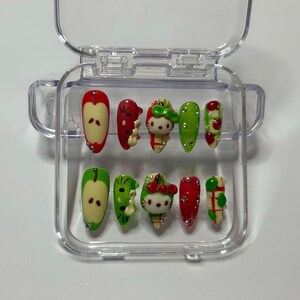 May include: A set of ten press-on nails in a clear plastic case. The nails feature various designs, including apples, cherries, and a Hello Kitty character, in red, green, and white. The nails are decorated with small, sparkling accents.