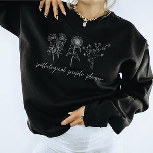May include: Black sweatshirt with white floral line art and the text "pathological people pleaser". The design features three different flower illustrations. The sweatshirt is a long-sleeved crew neck.