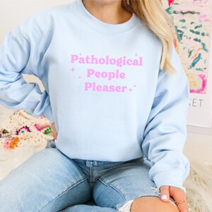 May include: A light blue sweatshirt with the text "Pathological People Pleaser" in pink lettering.