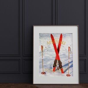 K&ouml;nnte beinhalten: Ein gerahmtes Poster mit einer Vintage-Skiszene. Das Poster zeigt zwei rote Ski, die im Schnee gekreuzt sind, mit Skist&ouml;cken auf beiden Seiten. Der Text "Apr&egrave;s Ski" ist in goldenen Lettern auf dem Poster gedruckt.