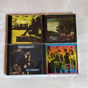 May include: Four compact disc cases with black spines and clear plastic fronts. The cases are arranged in a 2x2 grid. The top left case features a photo of Sting with the title "Sting Ten Summoner's Tales". The top right case features a photo of a tropical scene with the title "Fleetwood Mac Tango in the Night". The bottom left case features a photo of 10,000 Maniacs with the title "10,000 Maniacs MTV Unplugged". The bottom right case features a photo of a band with the title "The Best of Sting".