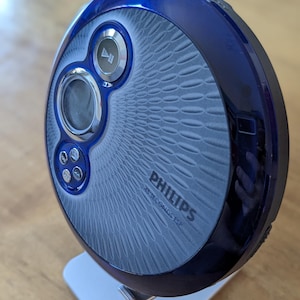 May include: A blue and silver Philips CD player with a textured design and a digital display. The player is on a white stand.