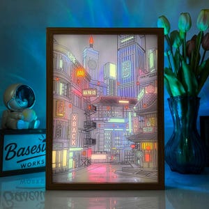 Könnte beinhalten: Ein rechteckiger Leuchtkasten mit einer detaillierten Illustration einer nächtlichen Stadtstraße mit Neonschildern, hohen Gebäuden und einer Straßenbahn. Das Bild ist in einen braunen Holzrahmen gefasst und strahlt ein sanftes Licht aus.