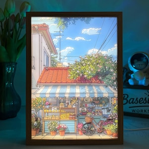 以下が含まれることがあります： 花屋、自転車、青い空のある魅力的な街並みを描いた、額入りの照明付きアートワーク。 木製フレームと鮮やかな色の詳細なイラストが特徴で、暖かく居心地の良い雰囲気を作り出しています。