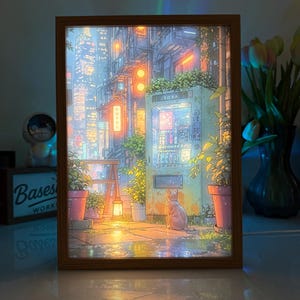 Puede incluir: Una obra de arte enmarcada e iluminada que representa una escena callejera japonesa con una máquina expendedora, un gato y edificios. La obra de arte está en un marco de madera y presenta colores cálidos. La palabra "SODA" es visible en la máquina expendedora.