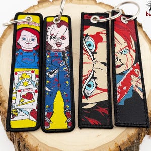 May include: Four fabric keychains featuring Chucky from the horror film franchise. The keychains depict Chucky in various poses, including a smiling doll and a bloody, menacing figure. The keychains are on a wooden slice.