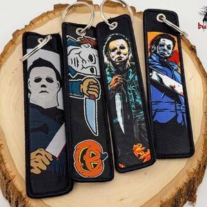 May include: Four black keychains with silver key rings, each featuring a different image of a horror movie character holding a knife. The keychains are displayed on a wooden slice. The images include a pumpkin and autumn leaves.