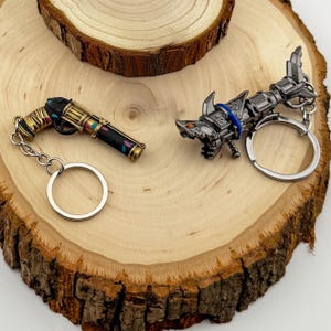 May include: Two metal keychains on a wooden surface. One is a gun-shaped keychain with gold and black accents, and the other is a shark-shaped keychain with silver and blue details. Both have key rings.