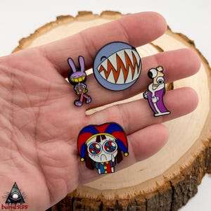 Puede incluir: Colecci&oacute;n de cinco pines de esmalte con personajes de dibujos animados. Los pines incluyen un conejo morado sonriente, un tibur&oacute;n con dientes naranjas, un personaje con una t&uacute;nica morada y un personaje triste con aspecto de payaso. Presentado sobre una superficie de madera.