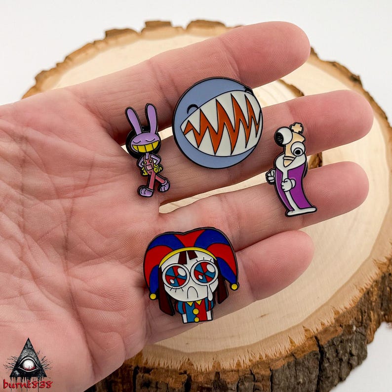 Puede incluir: Colecci&oacute;n de cinco pines de esmalte con personajes de dibujos animados. Los pines incluyen un conejo morado sonriente, un tibur&oacute;n con dientes naranjas, un personaje con una t&uacute;nica morada y un personaje triste con aspecto de payaso. Presentado sobre una superficie de madera.