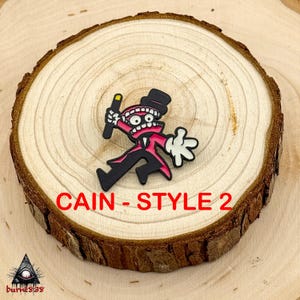 Puede incluir: Un pin de estilo caricatura con un personaje con traje negro, chaqueta rosa, sombrero de copa y guantes blancos, sosteniendo una varita con punta amarilla. El texto "CAIN - STYLE 2" se muestra en rojo. El pin est&aacute; sobre una rodaja de madera.