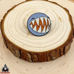 Puede incluir: Un pin redondo de esmalte con la cara de un tibur&oacute;n de dibujos animados. El pin tiene un fondo azul claro, dientes blancos y enc&iacute;as en zigzag naranjas. Se muestra sobre una rodaja de madera.
