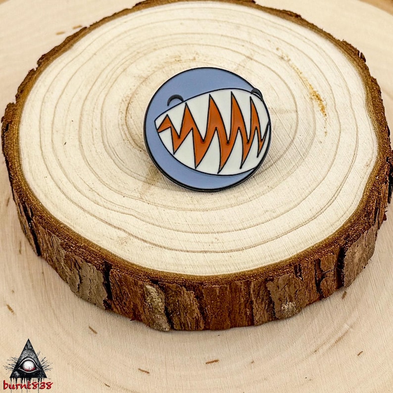 Puede incluir: Un pin redondo de esmalte con la cara de un tibur&oacute;n de dibujos animados. El pin tiene un fondo azul claro, dientes blancos y enc&iacute;as en zigzag naranjas. Se muestra sobre una rodaja de madera.