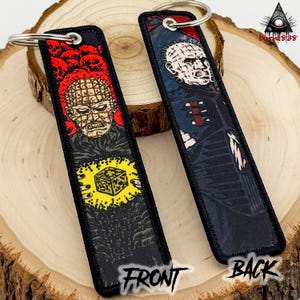 May include: Two black keychains with metal rings. One keychain features a red and black image of a character with pins in their head, and a yellow cube. The other keychain has a similar character in blue and black. The word "FRONT" and "BACK" are printed on the keychains.